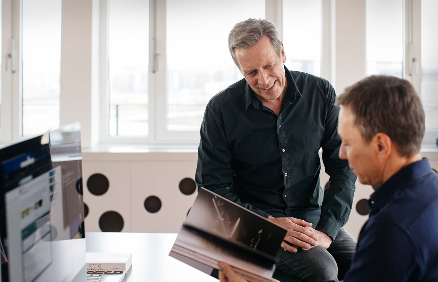 Zwei Männer im hellen Büro von BMP blättern gemeinsam in einem Magazin, links ein Bildschirmarbeitsplatz